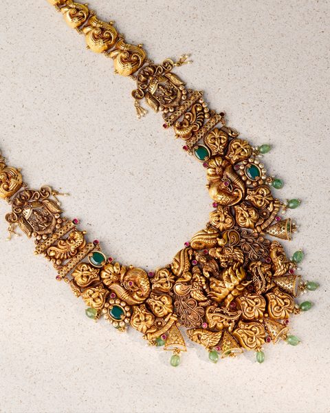 Long nakshi haram laid flat with peacock and deity plaques, emerald bead drops. Photographed at the shop.