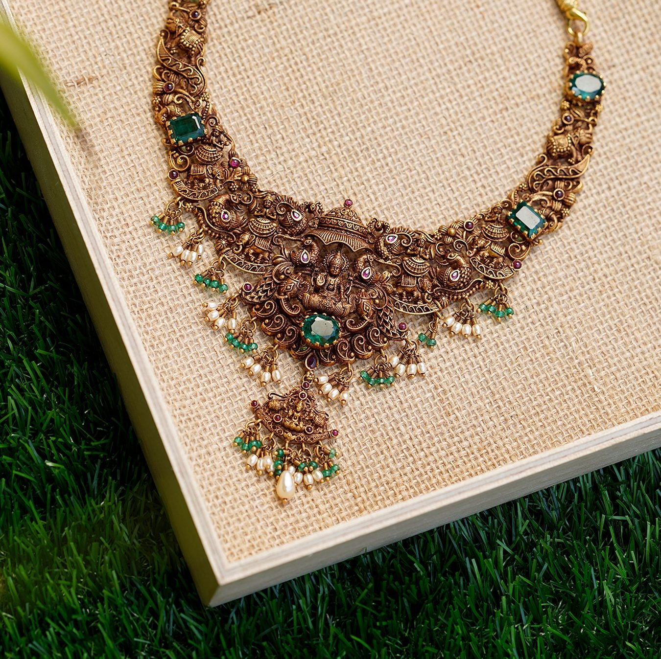 A nakshi necklace with emeralds and pearl drops laid on a jute board, photographed from above.