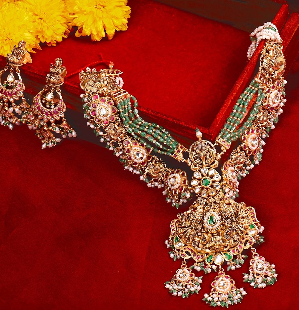 Emerald bead strands with a gold pendant and jhumkas beside yellow chrysanthemums.