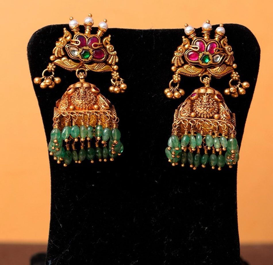 A pair of antique gold jhumkas with kempu, pearls and emerald bead drops on a black stand.