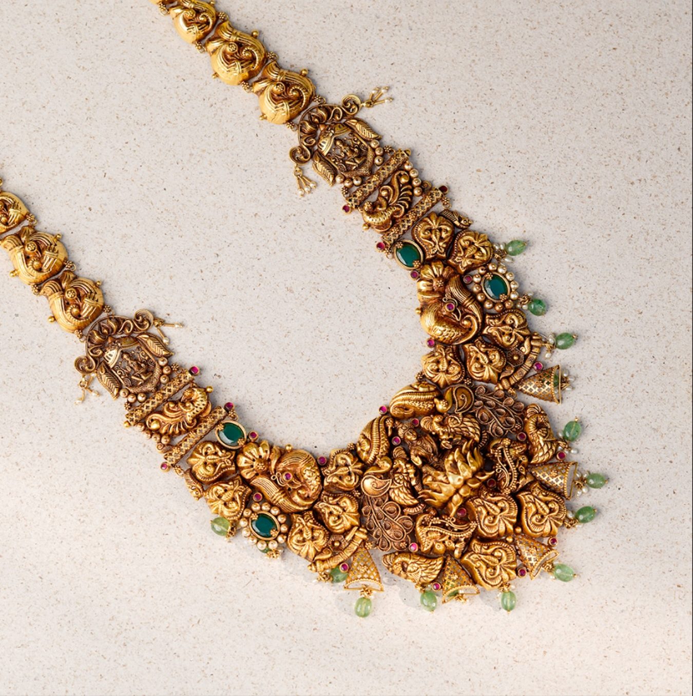 A long temple nakshi haram in antique gold with peacock and Lakshmi work and emerald bead drops, laid on a pale speckled stone counter, photographed from directly above.