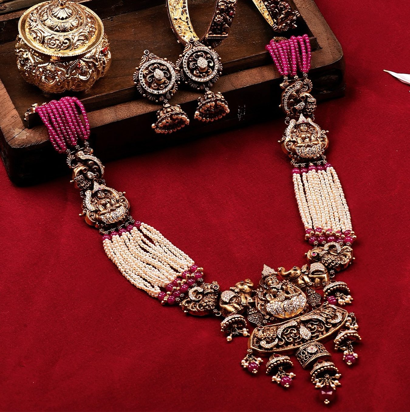 A bridal set laid out together on red velvet beside a carved wooden box: a long pearl and ruby haram with a Lakshmi pendant, two carved bangles, a pair of jhumkas and a kumkum box.